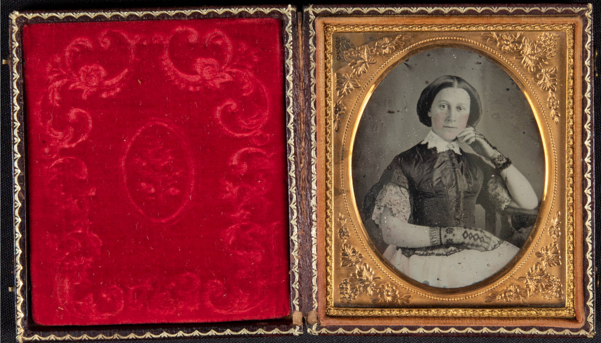 Black and white historic daguerreotype photograph inside of a red velvet lined case. Photograph is of a seated woman, her head resting aginst one hand propped on her lap. She wears gloves.